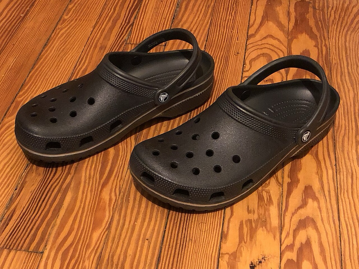 A pair of classic black Crocs foam clogs on a wooden floor, showing the signature ventilation holes and heel strap