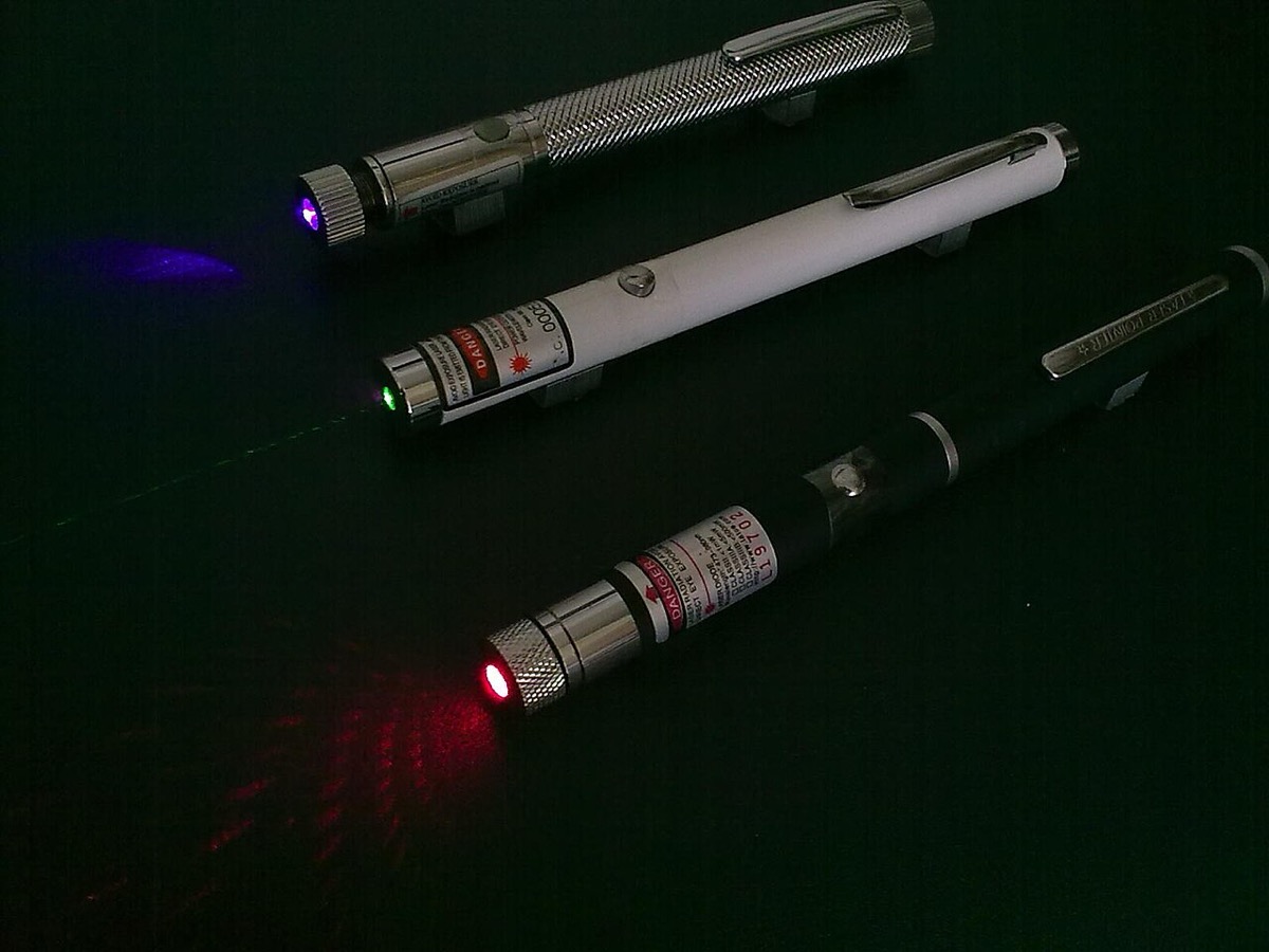 Three handheld laser pointers on a black background, each lit — a violet, a green, and a red beam and dot