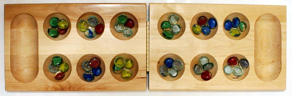 A wooden Mancala board with two rows of six round pits, each holding a scatter of colorful glass playing stones, and a large storage pit at each end