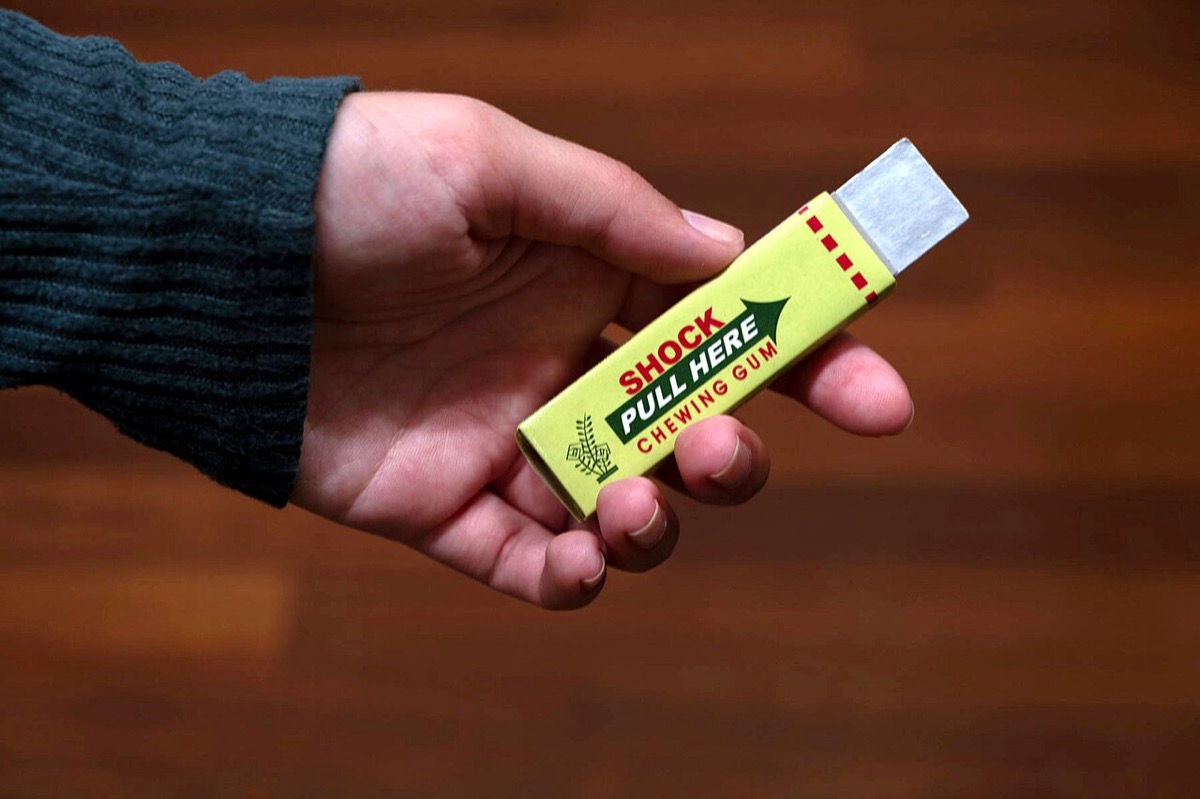 A hand offers a novelty shock chewing gum pack with one silver stick extended — pull the stick and it zaps you