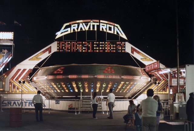 A Gravitron lit up at night on a carnival midway in the 1980s, its drum spinning while a small crowd waits at the gate