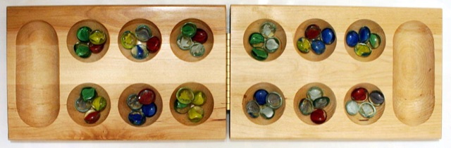 A wooden Mancala board with two rows of six round pits, each holding a scatter of colorful glass playing stones, and a large storage pit at each end