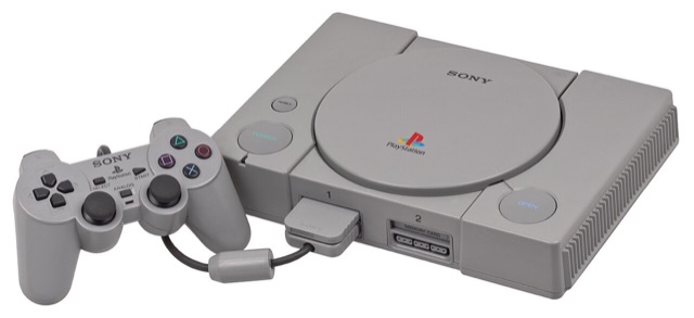A grey Sony PlayStation console shown with a DualShock controller and a memory card slotted into the front