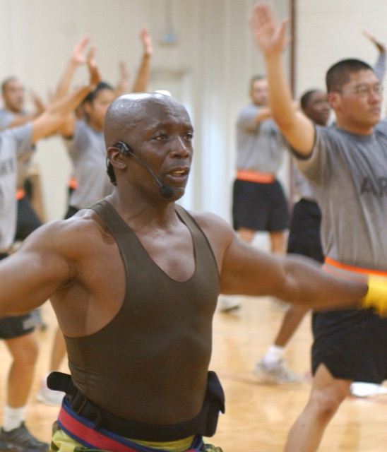Billy Blanks in a headset mic leading a Tae Bo class, arms out, with a room of people punching behind him