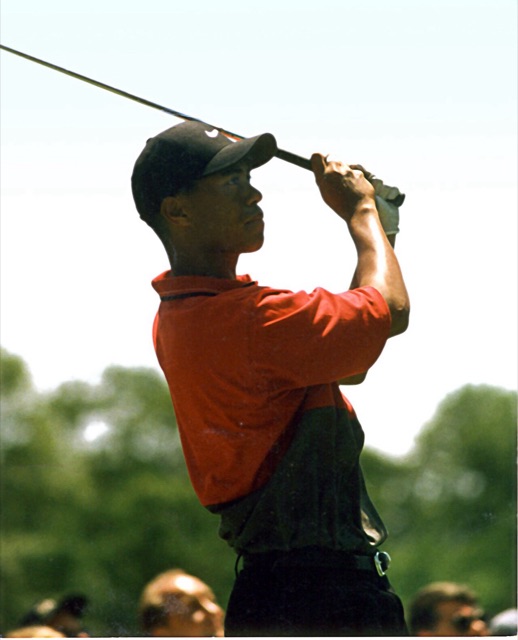 Tiger Woods in a red shirt and black Nike cap watching a tee shot, 1997