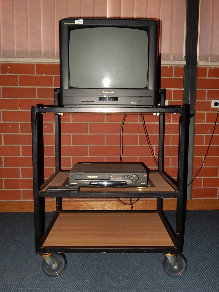 A CRT television strapped atop a tall black rolling AV cart, VCR and remote on the shelf below, against a school's brick wall